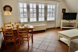 A dining area with a table set for four, a television on a stand, and a window with blinds. - Lindenhof Holiday Apartment No. 4 (Wenningstedt-Braderup)