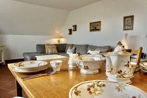 A dining area with a wooden table set with plates, bowls, and a pitcher. A gray sofa and a lamp are in the background. - Lindenhof Holiday Apartment No. 4 (Wenningstedt-Braderup)