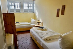 A small, compact bedroom with two single beds, a bedside table with a lamp, a window with blinds, and a door. - Lindenhof Holiday Apartment No. 4 (Wenningstedt-Braderup)