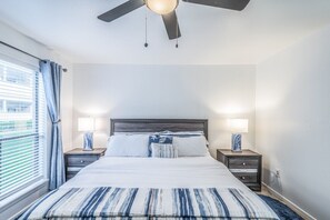 Une chambre à coucher comprenant un lit, deux tables de chevet, un ventilateur de plafond et une fenêtre avec des rideaux.