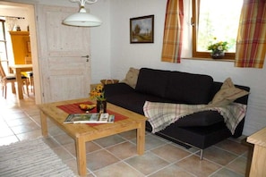Een woonkamer met een zwarte bank, een houten salontafel met tijdschriften, een vaas met bloemen en een raam met gordijnen.