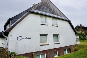 A two-story house with a white exterior and a dark roof. The house has three windows on the upper floor and a chimney. - House Cordula Holiday Apartment 38 (Wenningstedt-Braderup)