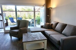 Ein Wohnzimmer mit Sofa, Sesseln und Couchtisch, eine Schiebetür zu einer Terrasse mit Sitzgelegenheiten.