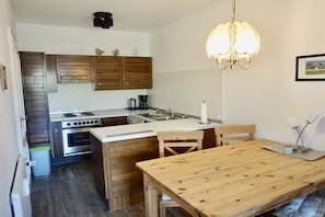 A compact kitchen with wooden cabinets, a stove, and a dining table with chairs. - House Cora Holiday Apartment 84 (Wenningstedt-Braderup)