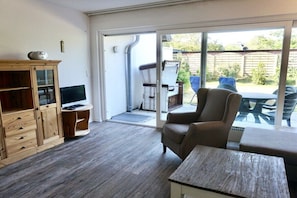 A room with a wooden cabinet, a television, a sofa, and a sliding glass door leading to an outdoor area with chairs and a table. - House Cora Holiday Apartment 84 (Wenningstedt-Braderup)