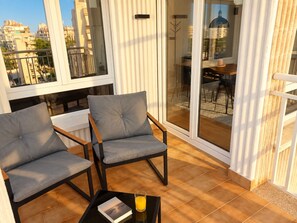 Een balkon met twee grijze stoelen, een kleine tafel met een boek en een glas sinaasappelsap, en een uitzicht op een stadsgezicht door grote glazen deuren.
