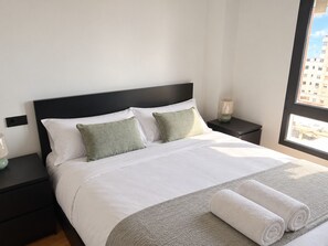 Un letto ben rifatto con lenzuola bianche e due cuscini, un tappeto grigio con trama decorativa e due asciugamani bianchi arrotolati sul letto.