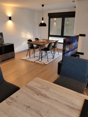 Een moderne woonkamer met een houten eettafel, zwarte stoelen, een blauwe bank, een televisie waarop Netflix wordt afgespeeld, en een groot raam met zonwering.
