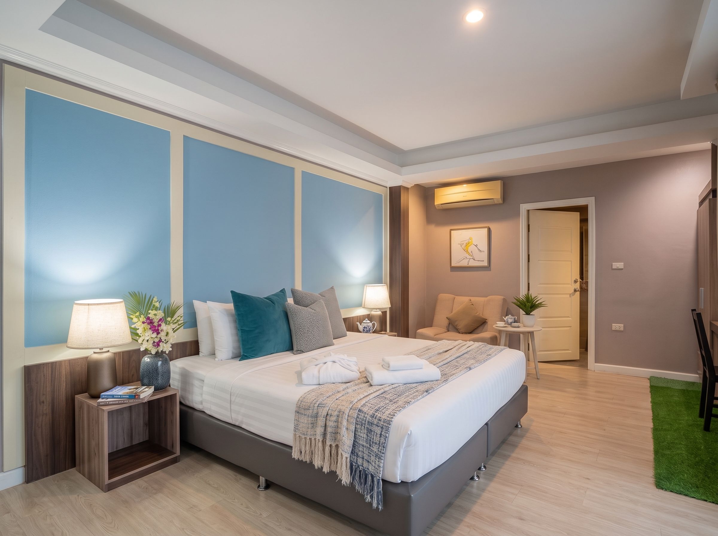 A modern hotel room with a large bed, bedside tables, a chair, a small table with a plant, and a door leading to another room.