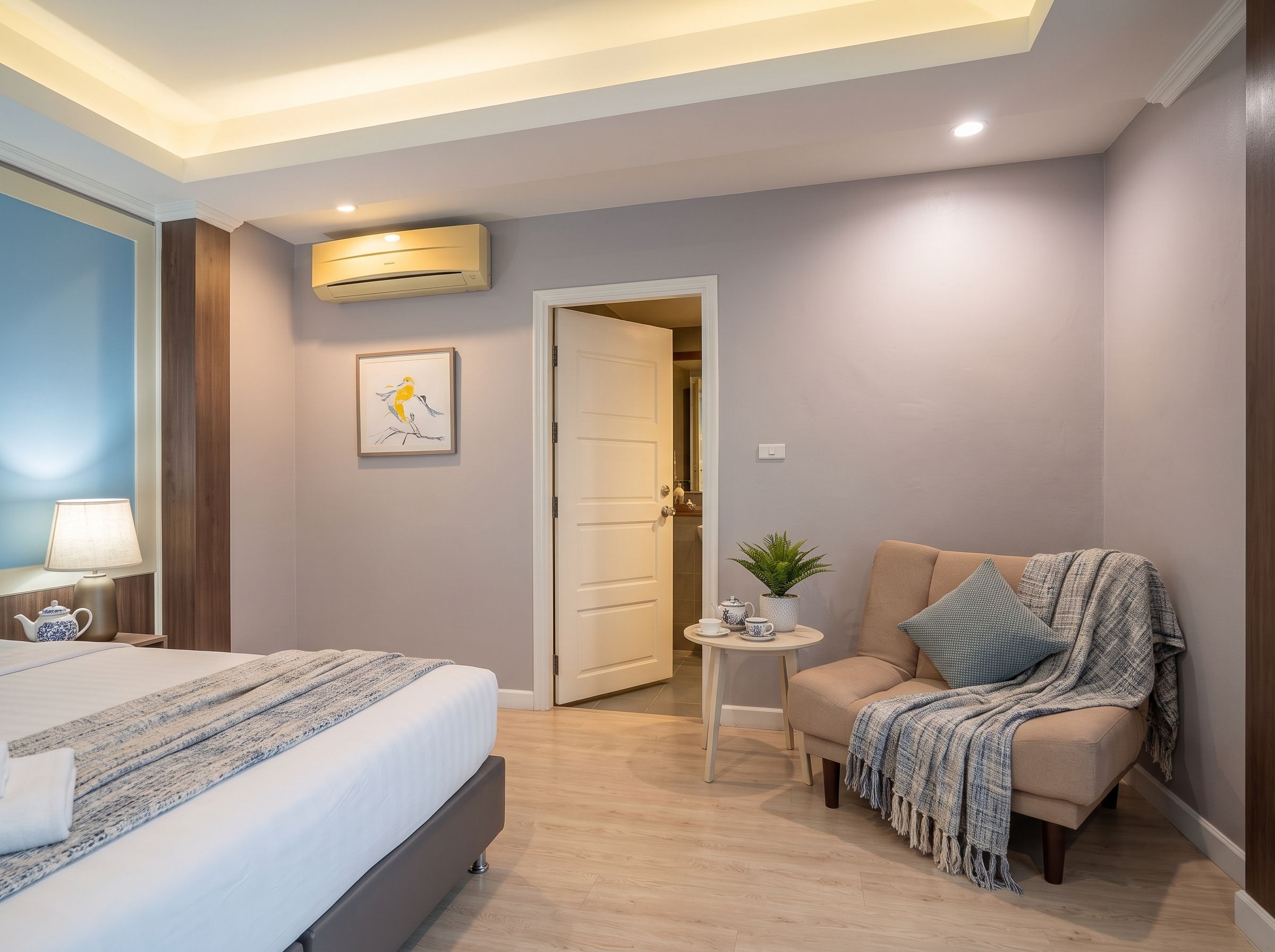 A hotel room with a bed, a chair, a small table with a plant, and a door leading to another room.