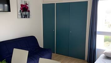 A room with a blue sofa, a glass table with chairs, a bed with pillows, teal-colored wardrobes, and a mounted TV.