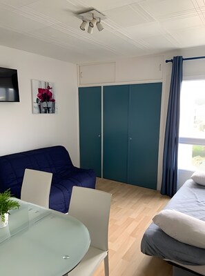 Een kamer met een blauwe bank, een glazen tafel met stoelen, een bed met kussens, blauwgroene kasten en een wandgemonteerde televisie.