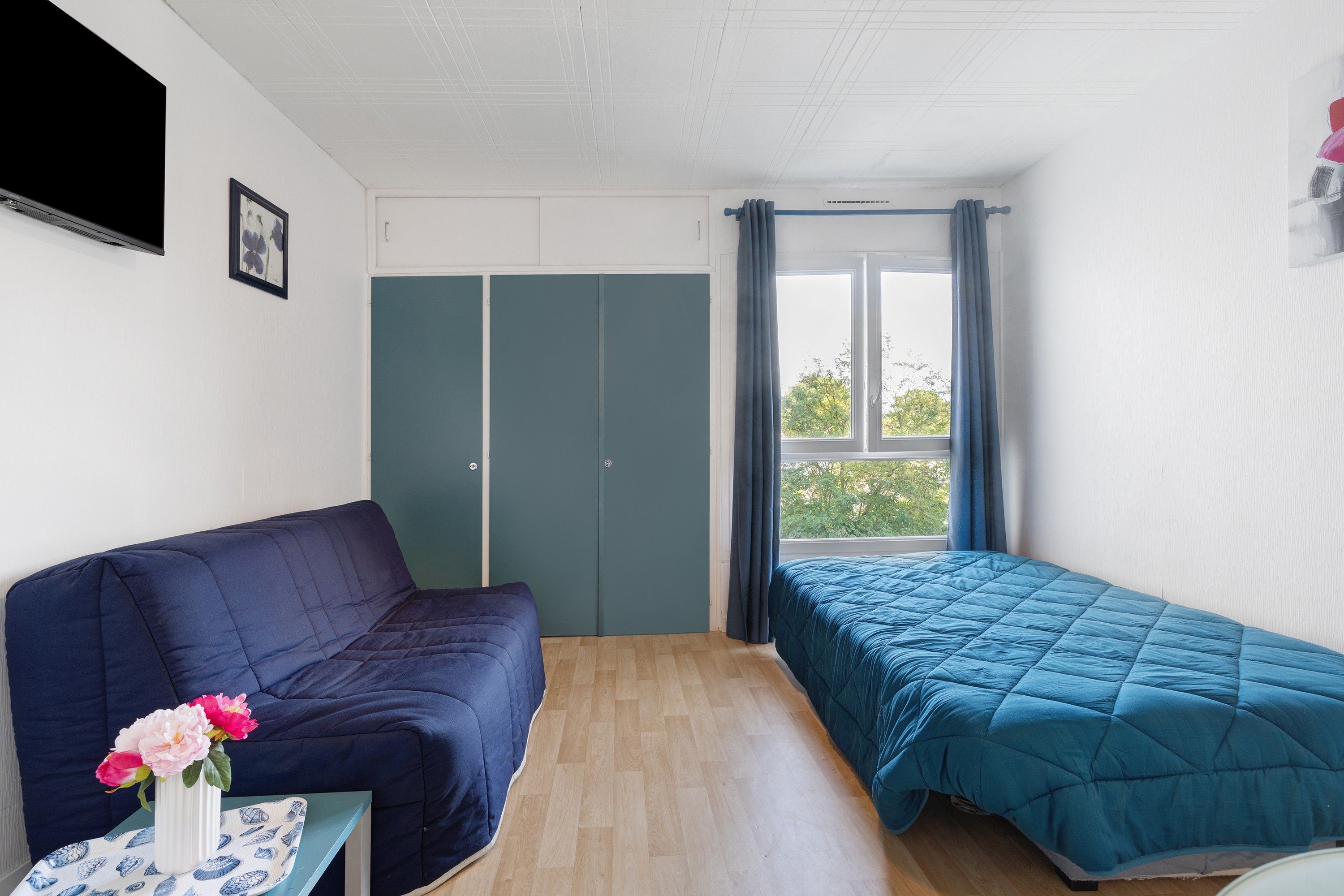 Ett rum med en säng, en soffa, ett litet bord med en vas med blommor och ett fönster med gardiner.