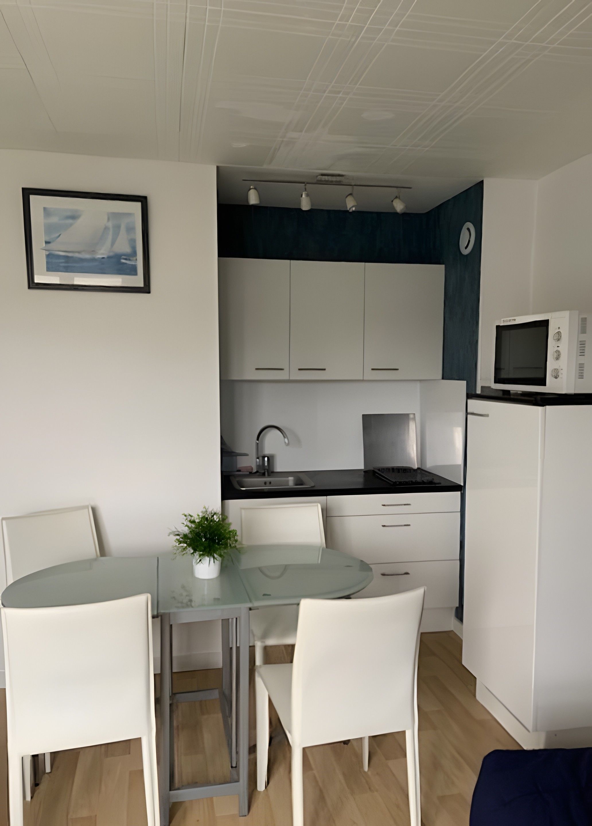 A compact kitchenette with a microwave, stove, and sink. There is a small dining area with a glass table and white chairs. A framed picture hangs on the wall.
