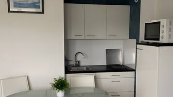 A compact kitchenette with a microwave, stove, and sink. There is a small dining area with a glass table and white chairs. A framed picture hangs on the wall.