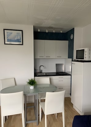 Een compact keukenhoekje met een magnetron, fornuis en spoelbak. Er is een klein eethoekje met een glazen tafel en witte stoelen. Een ingelijst schilderij hangt aan de muur.