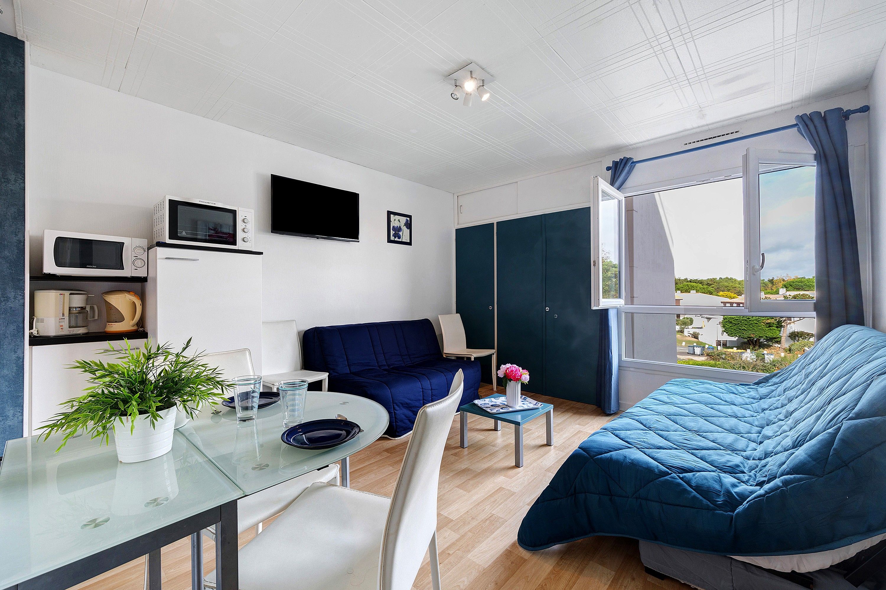 A compact living space with a bed, a seating area, a dining table, a microwave, a television, and a window with a view.