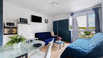 A compact living space with a bed, a seating area, a dining table, a microwave, a television, and a window with a view.