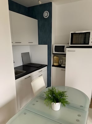 Een kleine keuken met witte kastjes, een zwart aanrecht, een magnetron, een koffiezetapparaat en een glazen tafel met een bloempot.