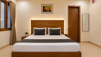 A hotel room with a large bed, a wooden headboard, a bedside table with a plant, a wall-mounted painting, and a door with a mirror.