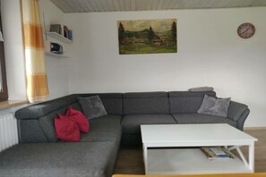 A living room with a grey sectional sofa, a white coffee table, a painting on the wall, a clock, and a radiator. - Ferienhof Waibel - Oberallgäu (Waltenhofen)