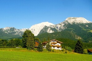 Ein Chalet in den Bergen mit einem Balkon, umgeben von üppigem Grün und blauem Himmel.