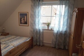 A bedroom with a single bed, a window with curtains, a radiator, and a small potted plant on the windowsill. - Holiday home with 2 bedrooms (Wangerland \/ Hooksiel)
