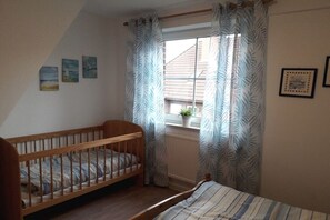 Een kamer met een houten wieg, een bed met gestreept beddengoed, een raam met gordijnen en een kleine potplant op de vensterbank.