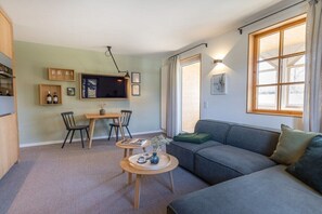 A modern living room with a grey sofa, a dining table with chairs, a TV mounted on the wall, and a large window with curtains. - Holiday apartment with 1 bedroom (Oberstdorf)