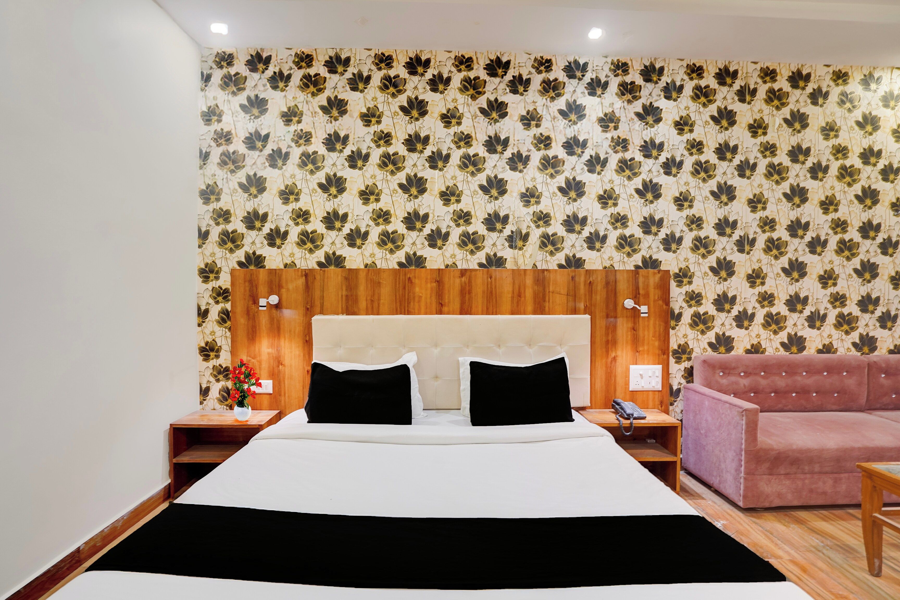 A hotel room with a bed, a wooden headboard, a pink sofa, and floral wallpaper.