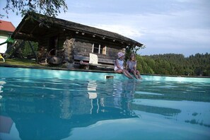 A wooden cabin with a stone foundation, a swimming pool, and a forested hill in the background. - Organic farm with 2 bedrooms (Missen-Wilhams)