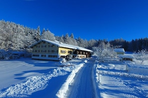 A snow-covered landscape with a building featuring a yellow facade and a brown roof, surrounded by trees and a clear sky. - Organic farm with 2 bedrooms (Missen-Wilhams)