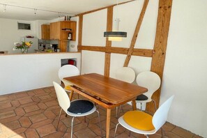 A dining area with a wooden table and white chairs, a kitchen in the background, and a hanging light fixture. - Holiday apartment with 1 bedroom (Lindau)