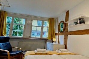 Ein Hotelzimmer mit einem Bett, einem Sessel, einem Fenster mit Vorhängen, einem Heizkörper und einem Regal mit Büchern.