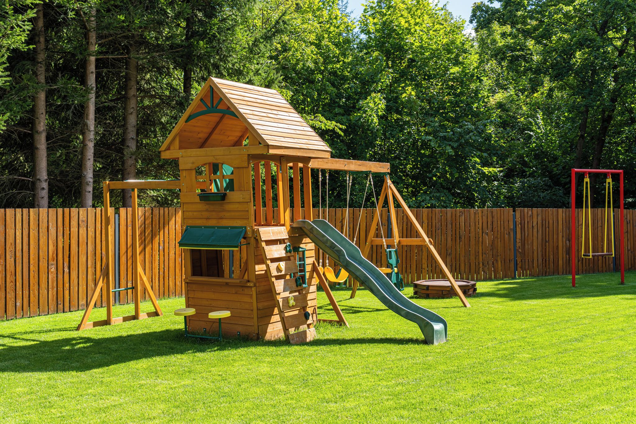 A wooden playhouse with a slide, surrounded by a wooden fence and trees.