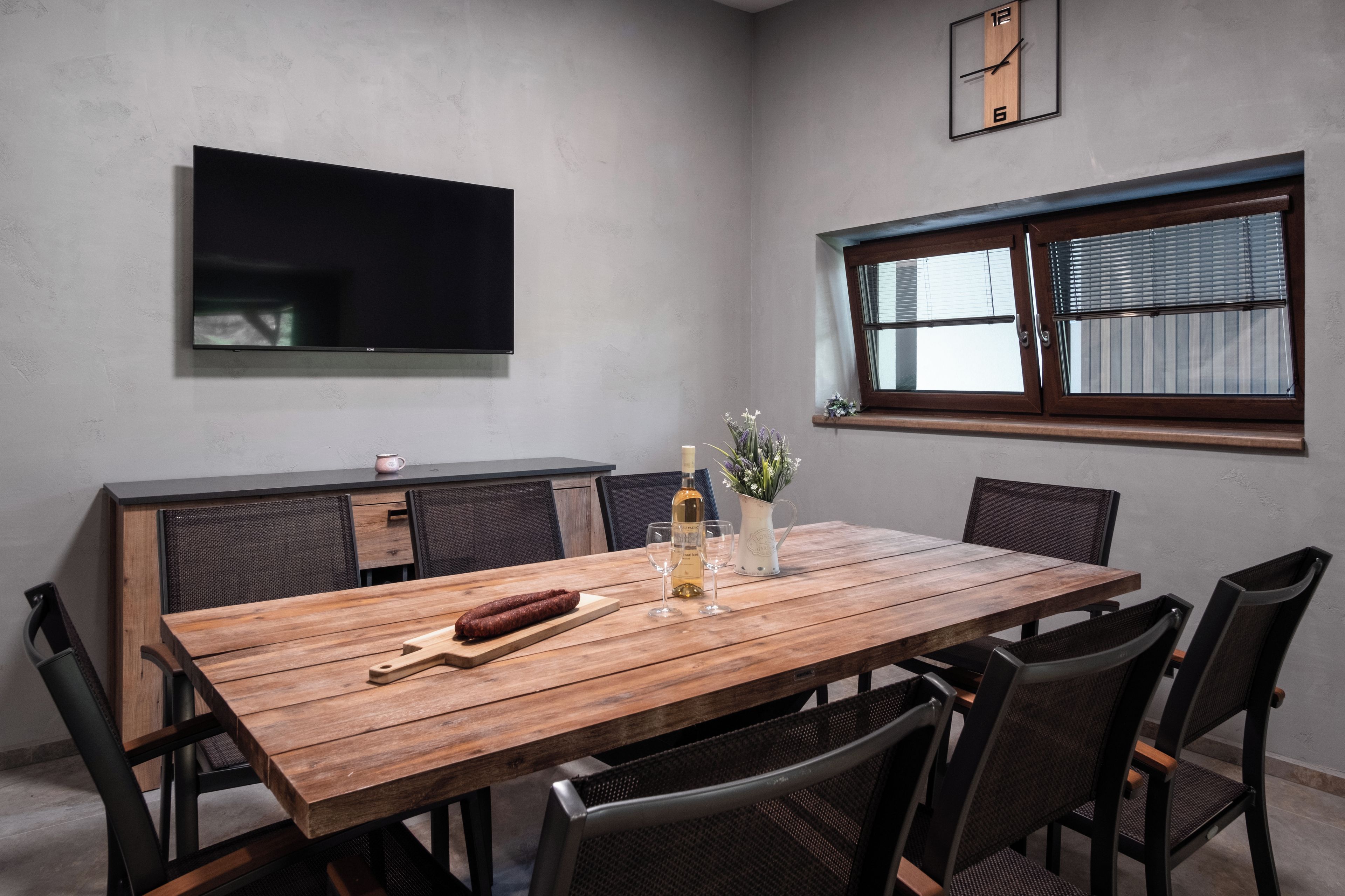 A dining area with a wooden table, chairs, a television mounted on the wall, and a window with blinds.