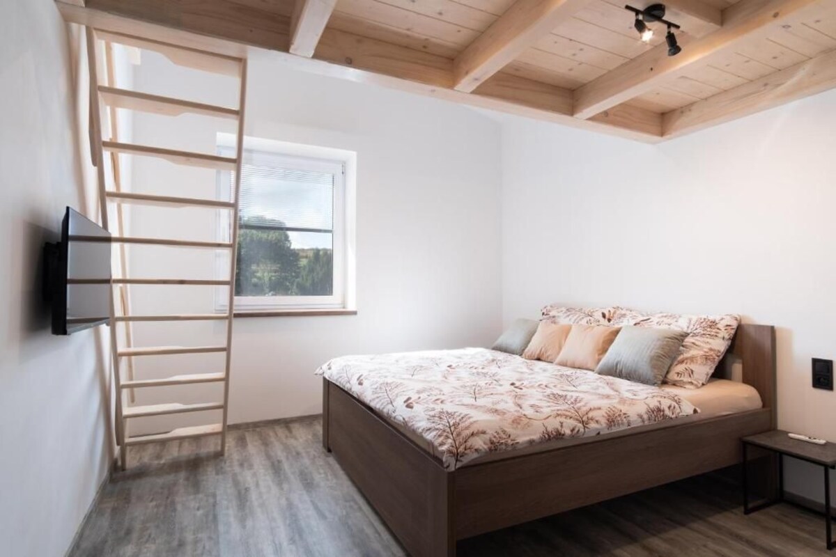 A bedroom with a bed, a TV mounted on the wall, a window, and a wooden ceiling.