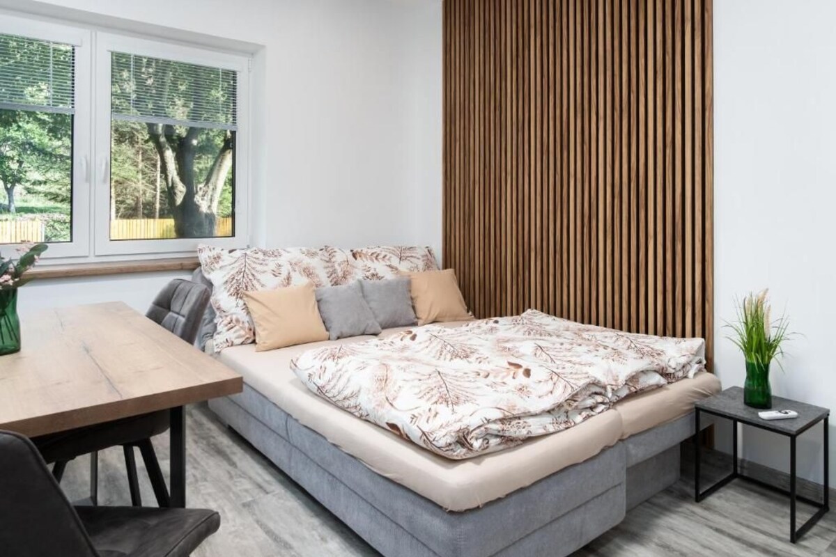 A modern bedroom with a bed, a wooden headboard, a window with blinds, a small table, and a plant.
