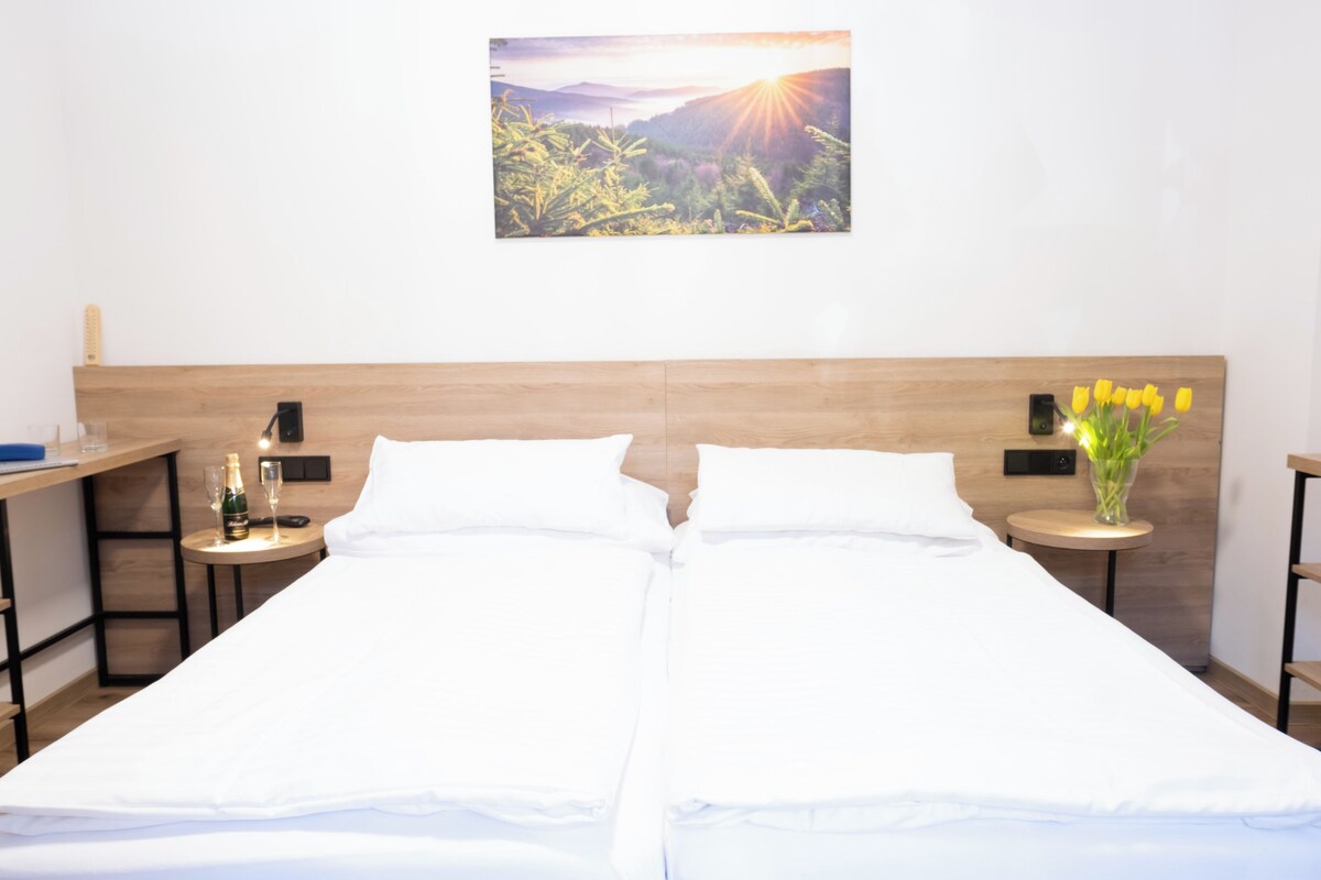 A double bed with white linens, a wooden headboard, two bedside tables with lamps, a nightstand with a bottle and glass, and a wall-mounted picture of a landscape.