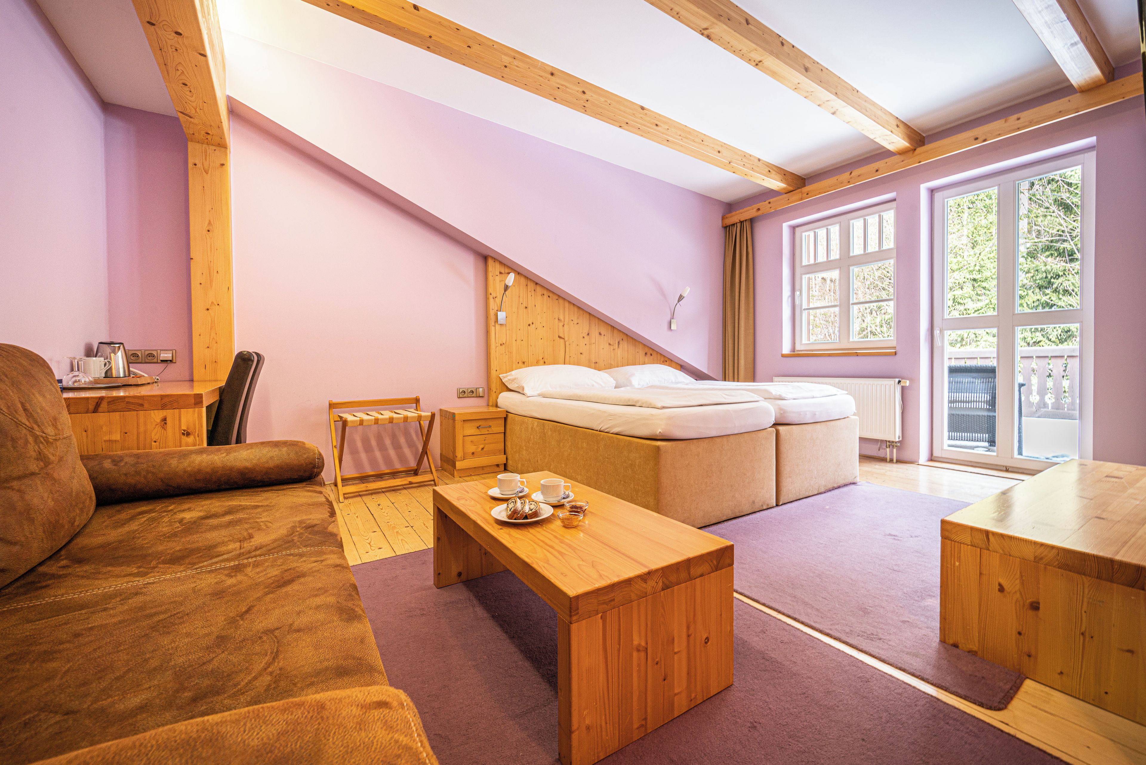 A cozy room with a wooden ceiling, a sofa, a small table with a tray, a bed with a headboard, and a window with curtains.