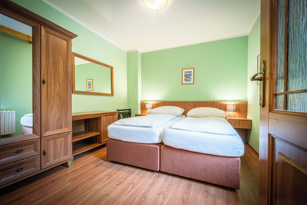 A room with a double bed, wooden wardrobe, bedside tables, a mirror, and a framed picture on the wall.