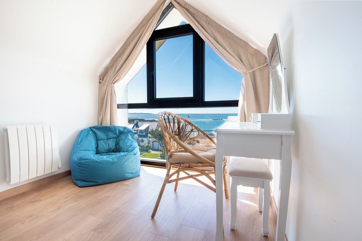 Una habitación con vista al mar, un escritorio blanco, un sillón de mimbre y un puff azul.