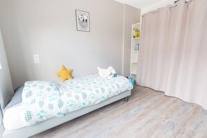 En säng med ett mönstrat överkast, ett litet sängbord, en nattlampa, en vägghylla med böcker och ett fönster med en gardin.