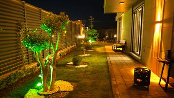 Espacio exterior nocturno con un sendero, plantas iluminadas, un banco y un contenedor de basura.