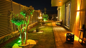 Zona exterior nocturna amb un camí, plantes il·luminades, una banca i un safataire.