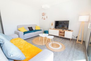Moderni olohuone, jossa on harmaa sohva, keltaisia ja sinisiä koristetyynyjä, litteänäyttöinen televisio, pieni valkoinen pöytä ja lattiavalaisin.