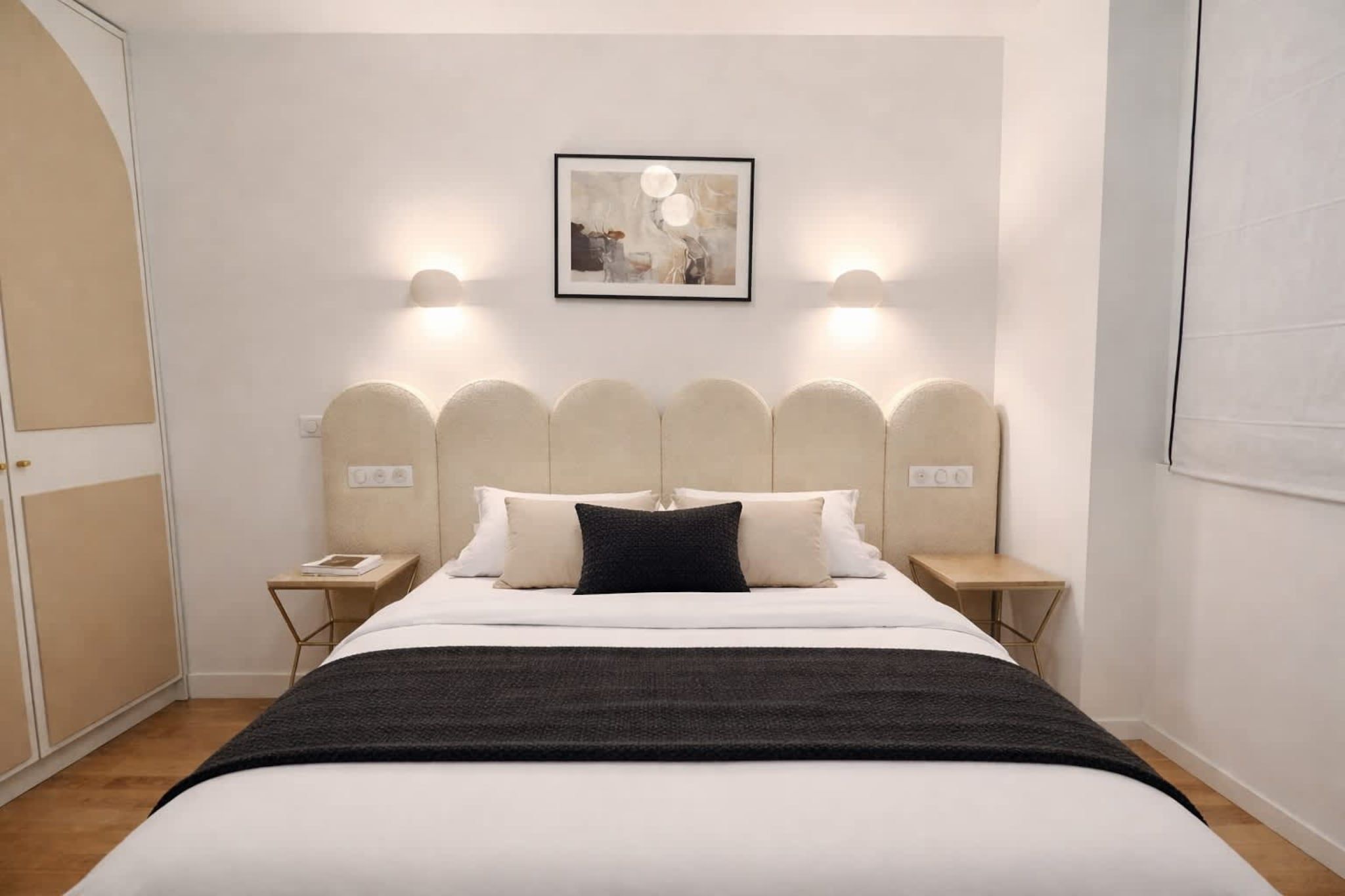 A hotel room with a bed, two bedside tables, a nightstand, a wall-mounted light fixture, and a framed picture above the bed.