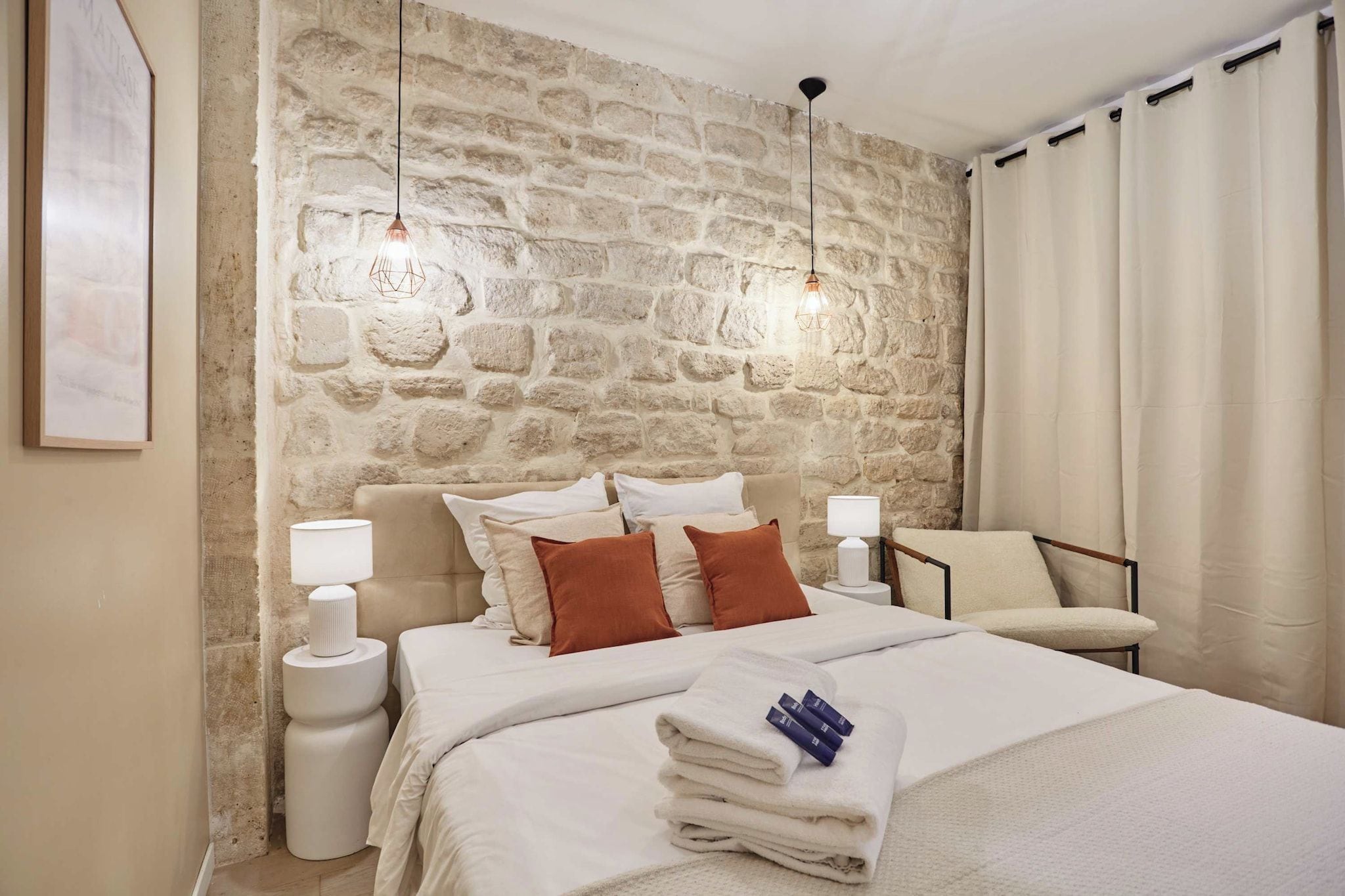 A hotel room with a stone wall, a bed with white bedding, two bedside lamps, a chair, and a framed picture on the wall.
