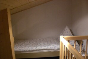 Ein kleines Zimmer mit einem Einzelbett, einem Holzkopfteil und einer schlichten Wand.