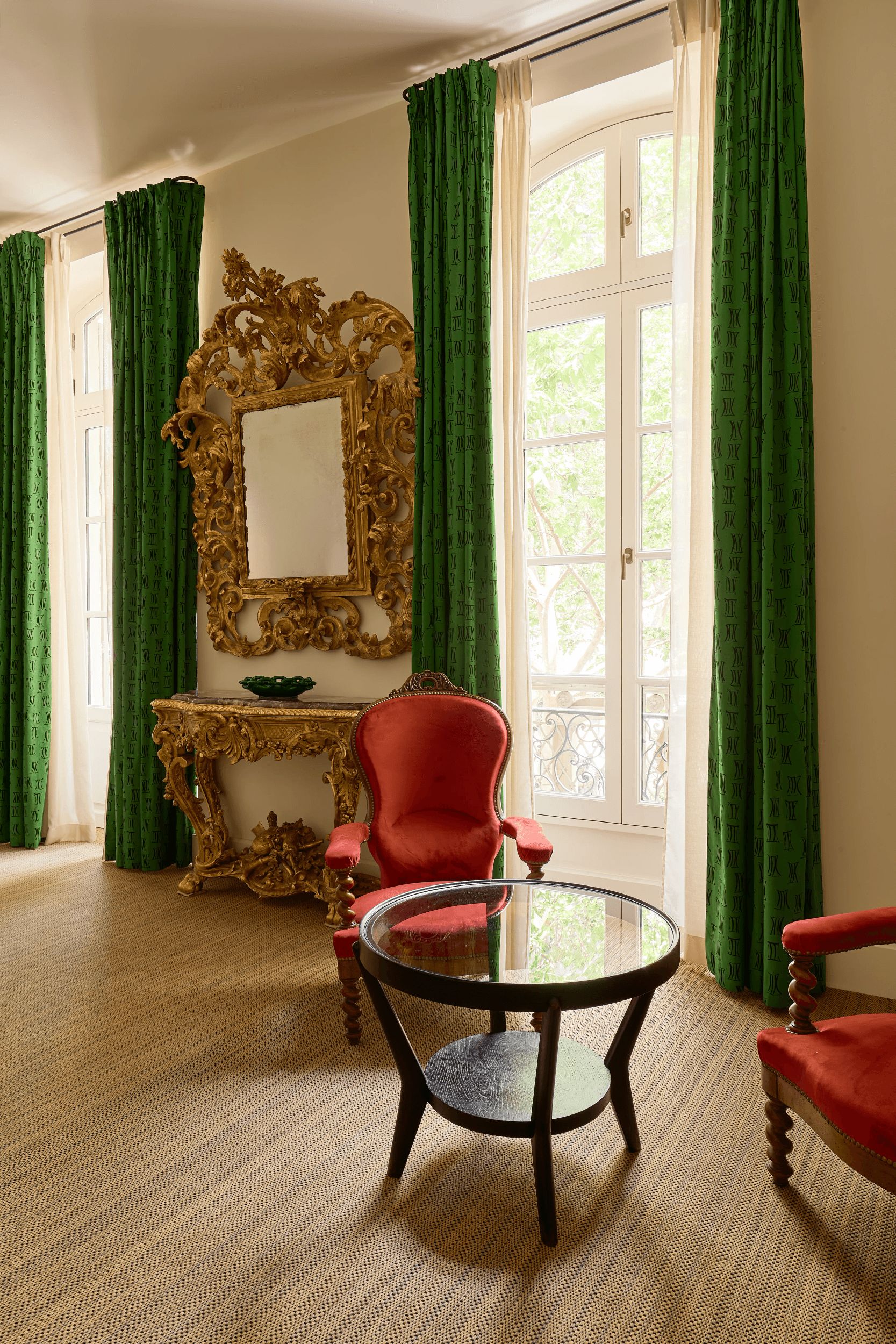 A room with a round table, a red chair, a gold-framed mirror, and green curtains.